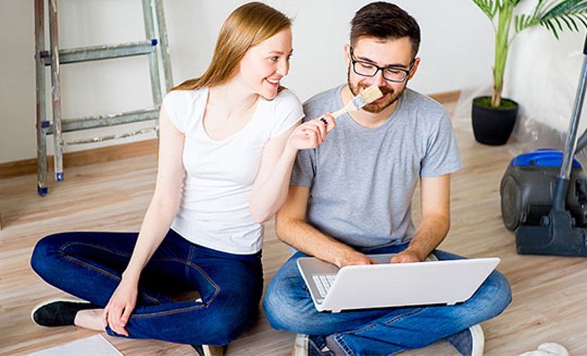 Renovierung - junges Paar sitzt mit Laptop und Pinsel auf Boden